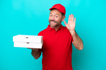 Pizza delivery man with work uniform picking up pizza boxes isolated on blue background saluting with hand with happy expression