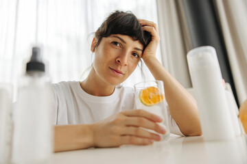 Fototapeta premium Calm young caucasian brunette woman looks at camera sitting at table in light room. Girl cleanses her body by drinking water with lemon. Health concept