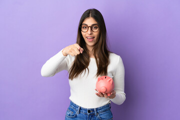 Young caucasian woman holding a piggybank isolated on purple background surprised and pointing front