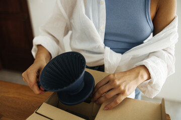 Close-up of fair-skinned woman taking out grey ceramic brew coffee cup from box indoors. Unboxing concept
