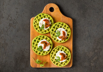 Round spinach waffles with cottage cheese, cherry tomatoes and parsley, on a wooden board. Dark gray background. Top view
