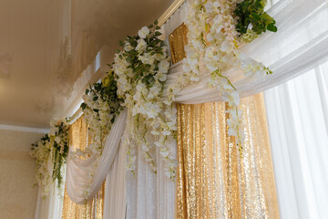 Windows decorated with white and golden shiny sparkling curtains, white artificial climbing flowers and green foliage. Reflection in glass ceiling