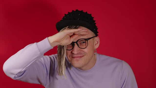 Young handsome man in black cap, glasses looking into distance, squinting. Blonde guy trying to see, holding hand over eyes on red background in studio.