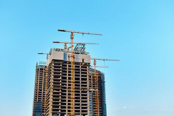 Three modern skyscrapers under construction with four yellow tower cranes along them in the process of installing windows on concrete structures against a clear blue sky in sunny weather