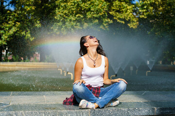 Caucasian young woman sitting by fountain enjoys coolness from water on sunny hot day, wearing...