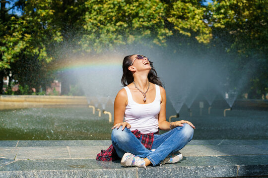 Caucasian young woman sitting by fountain enjoys coolness from water on sunny hot day, wearing casual clothes with sunglasses. Having fun against the backdrop of a rainbow from water grubs.