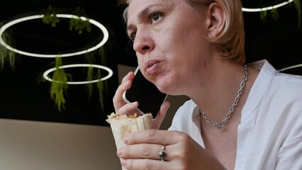a caucasoid woman with a wary expression chews a doner kebab and is silent while listening to her interlocutor on the phone during lunch in a restaurant.