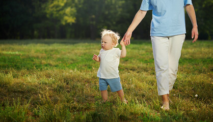 First baby steps. A young mother helps a baby boy walk on the grass in the park. Parental care and support