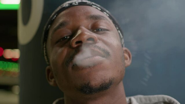 Portrait smoking black skin teenager smoking marijuana and exhaling smoke. African american man smokes cigarette inhaling harmful smoke with nicotine while standing near wall in an underpass.