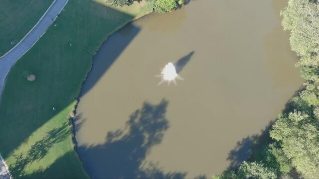 aerial footage of a silky green lake in the park with a water fountain surrounded by lush green trees and grass  at Lenox Park in Brookhaven Georgia USA