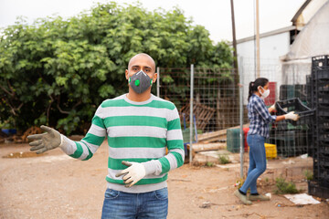 Naklejka premium Confident hispanic man farm worker in protective face mask standing outside near greenhouse buildings