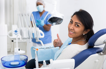 Happy latin american woman sitting in dental chair after teeth cure in modern clinic, gesturing...