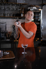 stylish young man hipster in orange t-shirt making mixing a cocktail in a dark loft cafe. alcohol drink in modern bar