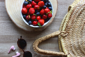 Round straw bag, brown sunglasses, pink earphones and bowl of berries on wooden table. Flat lay.