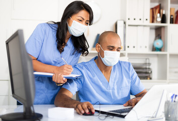 Fototapeta Professional doctor in surgical mask working in office with female assistant, filling up medical forms in laptop obraz