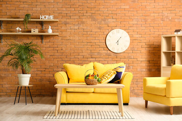 Interior of living room with sofa, armchair and table with fruit basket near brick wall