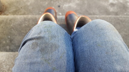 Fototapeta premium Point-of-view shot of grass-stained jeans and gumboots, woman is sitting on concrete stairs.