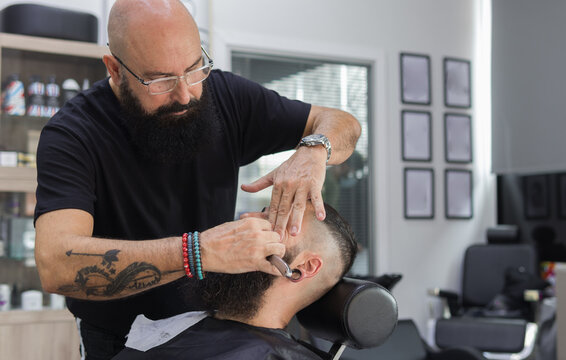 close up image of a tattooed hairdresser shaving a man's beard with a straight razor into a barbershop