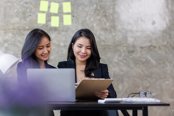 Cheerful asian businesspeople using a laptop in an office. Happy young entrepreneurs smiling while working together in a modern workspace. Two young businesspeople sitting together at a table.