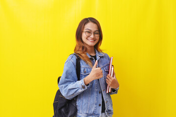 Young asian college student standing while holding books and showing thumbs up. Isolated on yellow