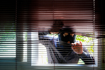 Unrecognizable Man Wearing Balaclava Face Mask Looking Through Venetian Blind. Keep your home...
