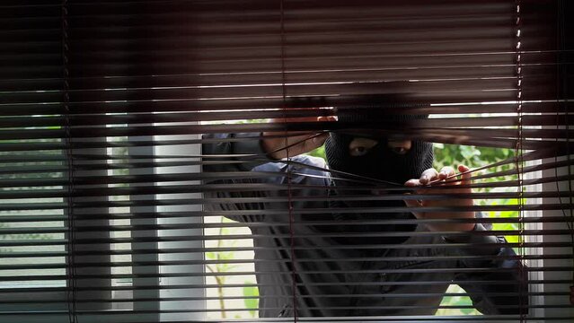 Unrecognizable Man Wearing Balaclava Face Mask Looking Through Venetian Blind. Keep your home secure during your summer holiday Concept.