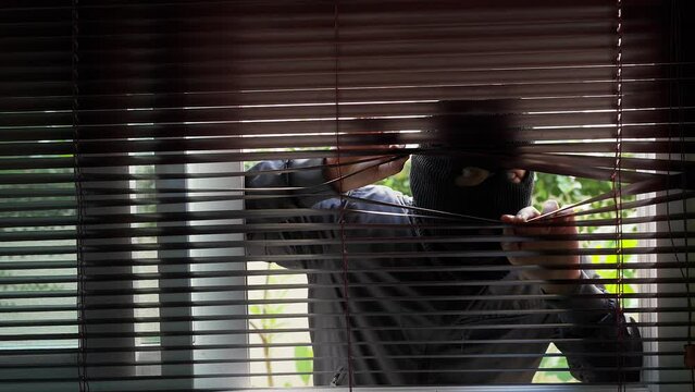 Unrecognizable Man Wearing Balaclava Face Mask Looking Through Venetian Blind.