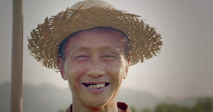 Slow-motion scene of a happy smiling Asian male farmer wearing a brown native shirt, an elderly native, standing in a plantation at sunset, lit up with an orange glow, behind is the mountains.
