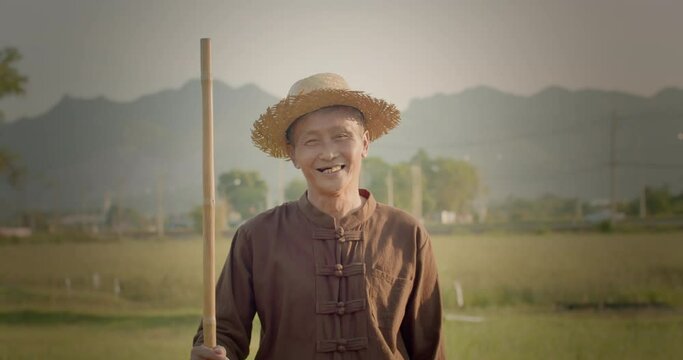 A happy smiling Asian male farmer wearing a brown native shirt, an elderly native, standing in a plantation at sunset, lit up with an orange glow, behind is the mountains.