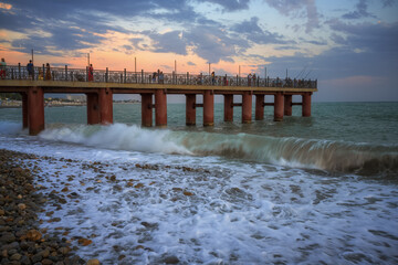 Evening walk along the old high pier over the sea in Adler (Russia) in summer. Waves crash against shore, forming foam