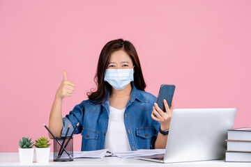 Young asian woman in casual clothes and wearing a mask to prevent covid 19 sit work at office desk with pc laptop isolated on pink background studio. She was holding a cell phone and gave a thumbs up.