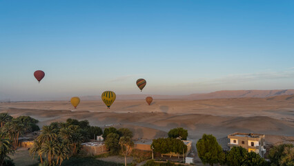 Bright balloons are flying over Luxor. Village houses and green vegetation are visible in the valley. In the distance - the sand dunes of the desert. Clear blue sky. Egypt.