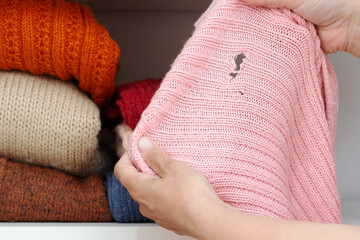 Cropped woman hands holding woolen knitted cloth with hole made by moth over wardrobe with stacks...