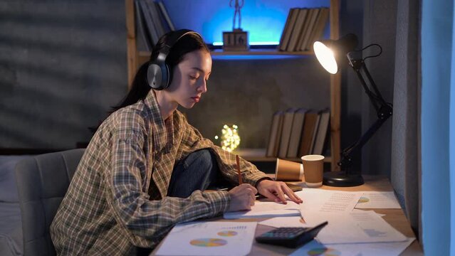 Asian student study hard in the evening. Using wearing headphones in a dark cozy room