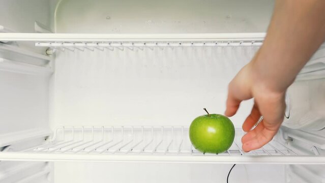 A man's hand opens an empty refrigerator and takes one green apple. Conceptual 4k raw video. Idea of ​​economic crisis, food shortage, poverty.