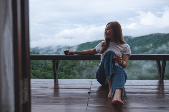 Portrait image of a woman sitting on wooden balcony with a beautiful mountains and nature view on foggy day