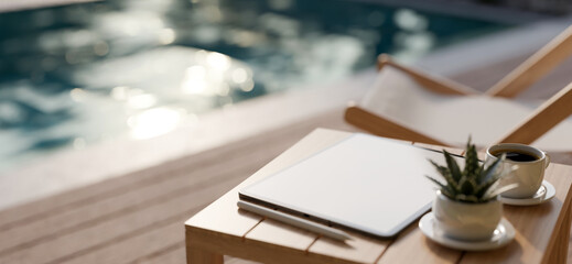 A portable tablet mockup is on a wooden outdoor side table over blurred swimming pool