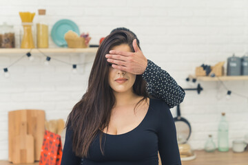 Unrecognizable man surprising his caucasian long-haired girlfriend with gift by covering her eyes with his hand. Kitchen interior. High quality photo