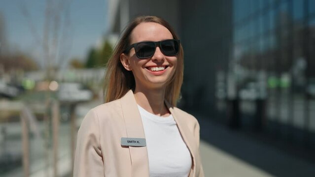 Portrait of attractive caucasian female worker in sunglasses looking at camera near business center. Smiling entrepreneur in formal jacket with badge posing at urban area.