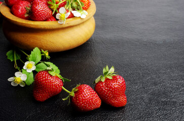Ripe strawberries in a wooden bowl with leaves and flowers on black. 