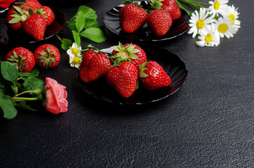 Ripe strawberries in black plates with flowers and leaves on a black table. 