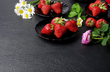 Ripe strawberries in black plates with flowers and leaves on a black table. 