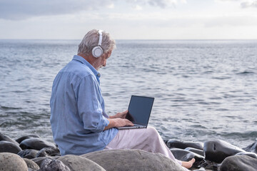 Senior active man sitting on the beach using laptop wearing headphones. Elderly bearded male barefoot enjoying sunset at sea, horizon over water