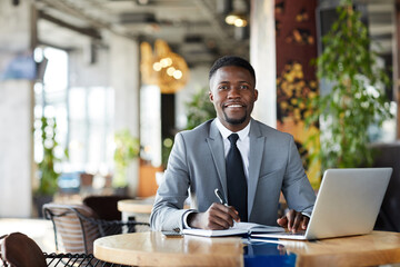 Smiling successful young African-American businessman with beard sitting at table in restaurant and making notes in organizer while planning timetable