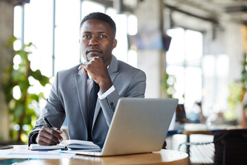Serious handsome young black businessman in stylish suit sitting at table with diary and laptop and looking at camera in restaurant
