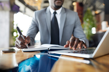 Close-up of black businessman in suit sitting at table in cafe and examining data on laptop and...