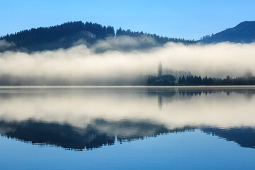 A blanket of mist reflected in the calm waters of a lake. Photographed at Lake Tutira, New Zealand