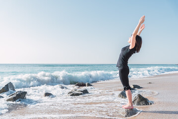 Side view of female stretching body and doing backbend while standing on rock in Tadasana. She is with raised arms on sea coast against blue sky during yoga session