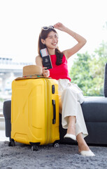 Fototapeta na wymiar Asian young happy cheerful female traveler in casual summer outfit with sunglasses sitting smiling on cozy sofa in living room holding passport and boarding pass ticket with yellow trolley luggage