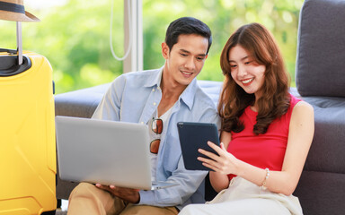 Asian young lovely boyfriend and girlfriend traveler couple sitting smiling on carpet in living room with trolley luggage while using laptop computer and tablet booking vacation summer trip online